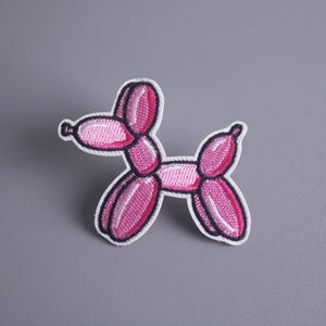 May include: Pink balloon animal shaped like a dog. The balloon animal is made of pink and white fabric and is embroidered with details.