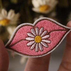 May include: Pink embroidered patch with a white daisy in the center. The daisy has a yellow center and white petals. The patch is shaped like a pair of lips.