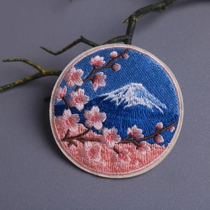 May include: Embroidered patch featuring a blue sky and a snow-capped mountain, with pink cherry blossoms. The round patch has a cream-colored border and is likely for clothing or accessories.