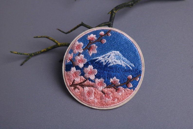 May include: Embroidered patch featuring a blue sky and a snow-capped mountain, with pink cherry blossoms. The round patch has a cream-colored border and is likely for clothing or accessories.