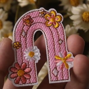 以下が含まれることがあります： 白と黄色のデイジーの花がついたピンクの刺繍された虹のパッチ。虹は白で縁取られており、質感のあるステッチのデザインになっています。