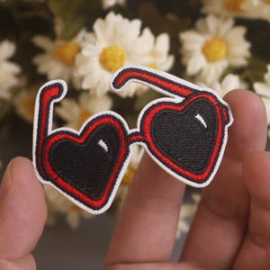 May include: Embroidered patch of heart-shaped sunglasses with black lenses and a red frame, outlined in white. The patch is held in front of a blurred background of white and yellow flowers.