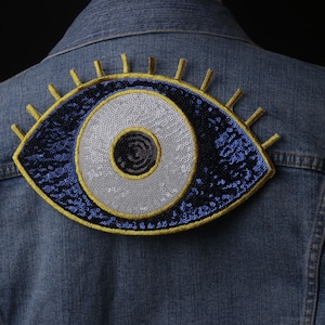 Könnte beinhalten: Ein blauer und weißer böser Blick-Patch mit goldenen Verzierungen, auf eine Jeansjacke genäht.