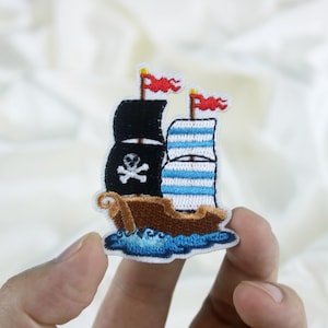 Könnte beinhalten: Ein gestickter Piratenschiff-Patch in Schwarzweiß mit einem Totenkopf und Knochen auf dem Segel. Das Schiff hat zwei rote Flaggen und segelt auf blauen Wellen.