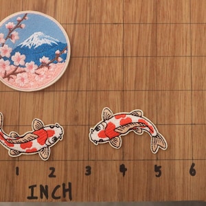 May include: Embroidered patches featuring Japanese motifs. One patch depicts Mount Fuji with cherry blossoms, and two others show koi fish. The patches are on a wooden surface with inch measurements.