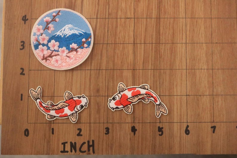 May include: Embroidered patches featuring Japanese motifs. One patch depicts Mount Fuji with cherry blossoms, and two others show koi fish. The patches are on a wooden surface with inch measurements.