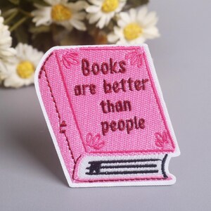 May include: A pink embroidered patch in the shape of a book. The patch features the text "Books are better than people" in dark pink thread. The book has a white border and decorative floral accents.
