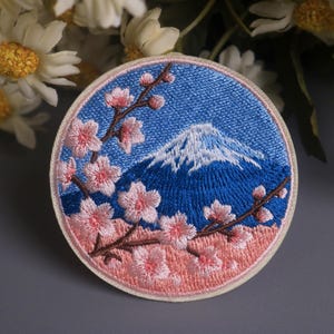 May include: Embroidered patch featuring a blue sky and a snow-capped mountain, with pink cherry blossoms on brown branches. The patch has a light pink border and a blue background.