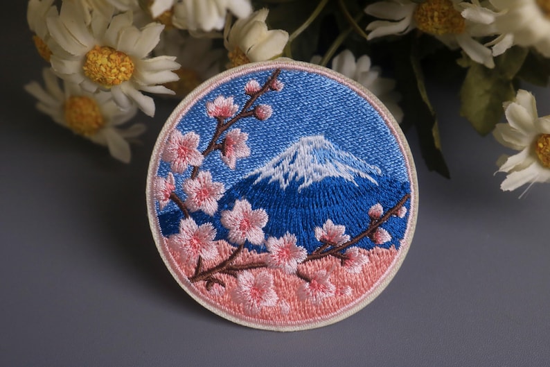 May include: Embroidered patch featuring a blue sky and a snow-capped mountain, with pink cherry blossoms on brown branches. The patch has a light pink border and a blue background.