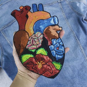 May include: A colorful heart-shaped patch with sequins, featuring a detailed anatomical design of the human heart. The patch is made of fabric and has a black background with red, blue, green, and pink sequins.