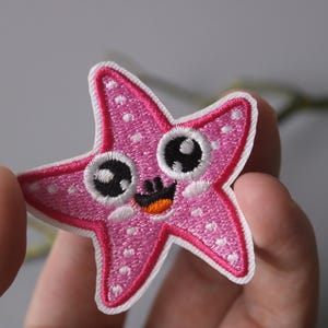 Puede incluir: Una estrella de mar bordada de color rosa y blanco con una cara sonriente y ojos grandes. La estrella de mar es un parche que se puede planchar sobre la tela.