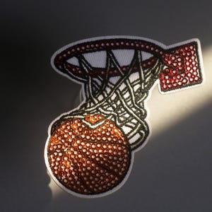 Peut inclure: &Eacute;cusson brod&eacute; repr&eacute;sentant un ballon de basket passant dans un panier. Le patch a un fond blanc avec un design rouge et noir. Le ballon est marron avec des coutures blanches, et le filet est noir.