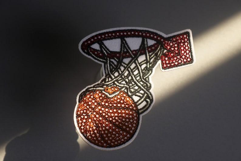 Peut inclure: &Eacute;cusson brod&eacute; repr&eacute;sentant un ballon de basket passant dans un panier. Le patch a un fond blanc avec un design rouge et noir. Le ballon est marron avec des coutures blanches, et le filet est noir.