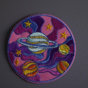 May include: A pink, purple, and blue embroidered patch with a planet and stars. The planet has a ring around it and is surrounded by swirling clouds.