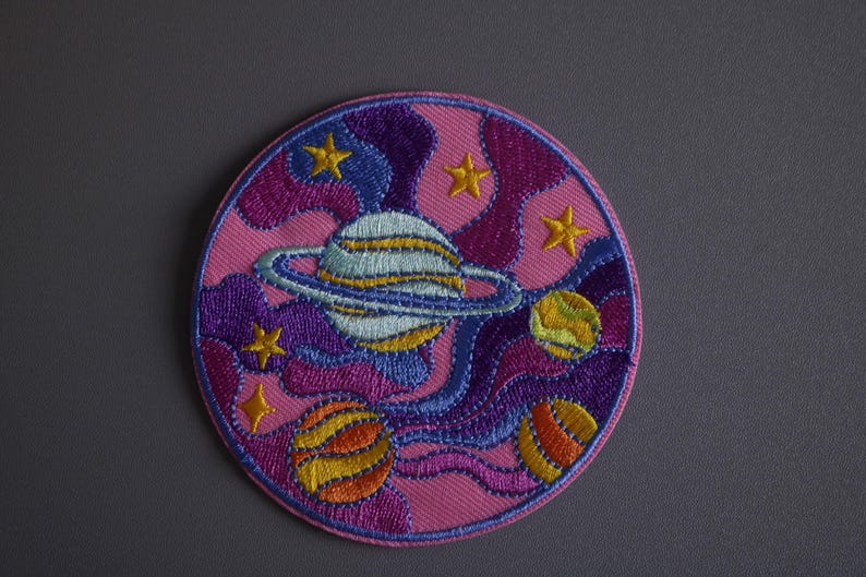 May include: A pink, purple, and blue embroidered patch with a planet and stars. The planet has a ring around it and is surrounded by swirling clouds.