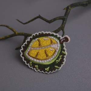 Pode incluir: Um remendo bordado que representa uma fruta de durian, com pontos amarelos e verdes. O remendo é contornado de branco e tem um caule castanho.
