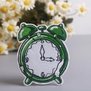 May include: Embroidered patch featuring a green alarm clock design. The clock face is white with black numerals and hands pointing to 3 o'clock. The patch has a green outline and details, with a white background.