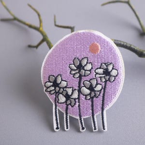 May include: A purple embroidered patch with five white flowers with black outlines and a small pink circle in the background.