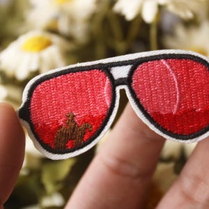 Puede incluir: Parche bordado rojo en forma de gafas de sol con una silueta marrón de un camello en las lentes.