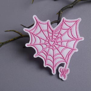 May include: Embroidered patch in the shape of a heart-shaped spiderweb with a small heart in the center. The web is pink, with a small pink spider at the bottom. The patch is on a white background.