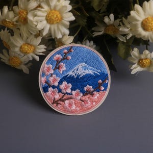 May include: Embroidered patch featuring Mount Fuji with a snow-capped peak, set against a blue sky. Pink cherry blossoms frame the mountain, with a light pink border. The patch is round and has a denim-like texture.