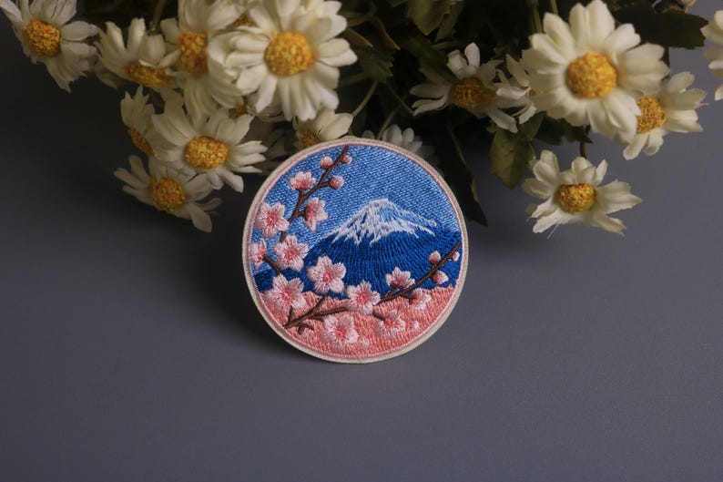 May include: Embroidered patch featuring Mount Fuji with a snow-capped peak, set against a blue sky. Pink cherry blossoms frame the mountain, with a light pink border. The patch is round and has a denim-like texture.