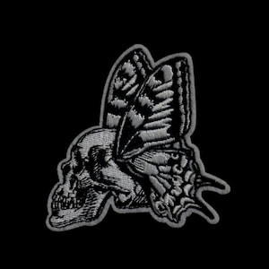 Könnte beinhalten: Ein gestickter Patch in Schwarzweiß, der einen Schädel mit einem Schmetterling darauf zeigt.