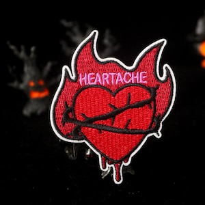 Può includere: Toppa ricamata con un cuore rosso trafitto da una spina nera, circondato da fiamme. La parola "HEARTACHE" è ricamata in rosa. La toppa ha un bordo bianco e un design di sangue che gocciola.