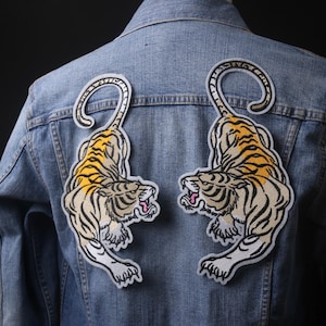 May include: Two embroidered tiger patches on a blue denim jacket. The tigers are facing each other with their mouths open and their tails curled around their bodies. The tigers are a light brown color with yellow stripes.