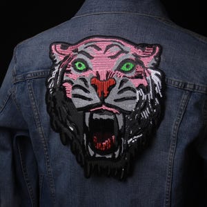 Puede incluir: Un parche de cabeza de tigre de lentejuelas rosa y negro. El tigre tiene ojos verdes y una expresión feroz.