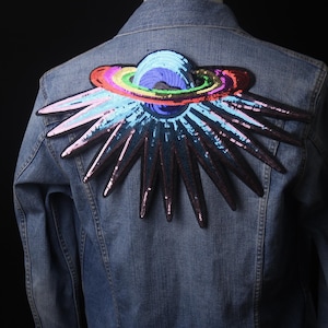 Può includere: Una giacca di jeans con una grande toppa di un pianeta colorata e scintillante, con un anello arcobaleno e un design a forma di stella.