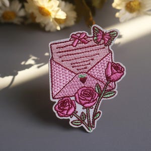 Peut inclure: Écusson brodé représentant une enveloppe rose avec un nœud et un papillon, entourée de roses roses. L'écusson a une bordure blanche et est destiné aux vêtements ou accessoires.
