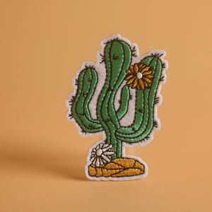 May include: Embroidered cactus patch with green and black stitching, featuring a yellow and white flower. The patch has a white border and is set against a light orange background. The cactus design is a fun, decorative element.