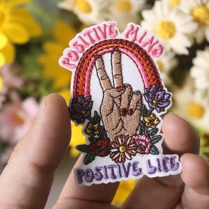 May include: A white embroidered patch with a rainbow, peace sign, and flowers. The text "Positive Mind" is above the peace sign and "Positive Life" is below.