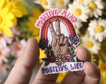 May include: A white embroidered patch with a rainbow, peace sign, and flowers. The text "Positive Mind" is above the peace sign and "Positive Life" is below.