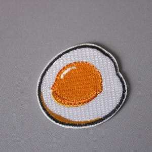 May include: A white embroidered patch with a yellow yolk in the center, depicting a fried egg.