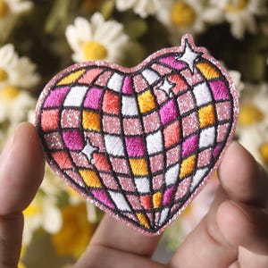 May include: A pink heart-shaped embroidered patch with a disco ball design. The disco ball is made up of pink, orange, white, and black squares. There are three white stars on the patch.