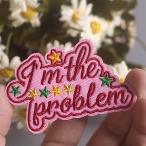 Può includere: Toppa in tessuto rosa con il testo "I'm the problem" in ricamo rosso. La toppa è decorata con cinque piccole stelle ricamate in verde e giallo.