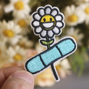 May include: Embroidered iron-on patch featuring a white daisy with a yellow smiley face and a blue bandage stem.