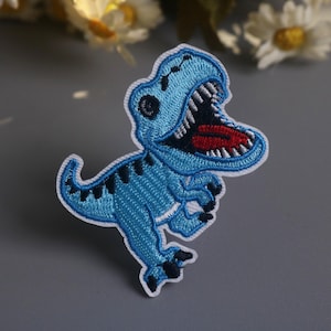 Op de afbeelding: Geborduurde patch met een blauwe dinosaurus met een open mond, met witte tanden en een rode tong. De dinosaurus heeft zwarte accenten en een witte rand. Geschikt voor kleding en accessoires.