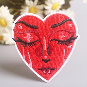May include: Embroidered patch in the shape of a red heart with a face. The heart has closed eyes with long black eyelashes, a small nose, and a downturned mouth. Tears are embroidered below the eyes. The patch has a white border.