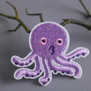 Puede incluir: Parche de pulpo bordado de color morado con contorno blanco y ojos rosas.