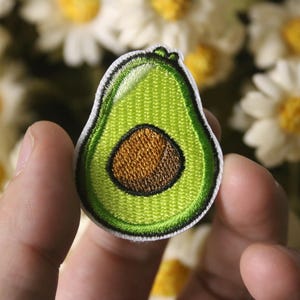 Könnte beinhalten: Ein grüner Avocado-Stickerei-Aufnäher mit einem braunen Kern. Der Aufnäher wird in einer Hand vor einem Hintergrund aus weißen Gänseblümchen gehalten.