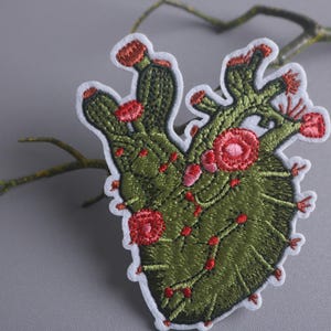 Könnte beinhalten: Grüner gestickter Kaktus-Herz-Patch mit roten Blüten. Der Patch befindet sich auf einem grauen Hintergrund.