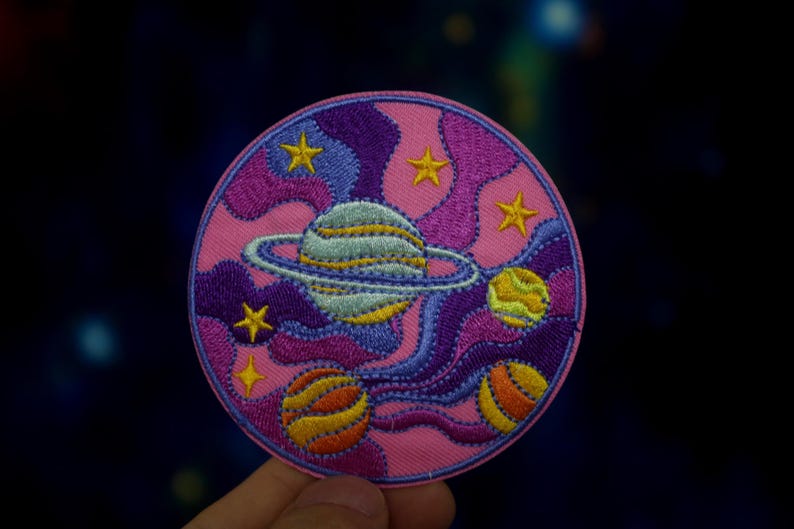May include: A pink, purple, and blue embroidered patch with a planet, stars, and swirling clouds. The planet has a ring around it and is light blue with yellow stripes. The stars are yellow and the clouds are purple.