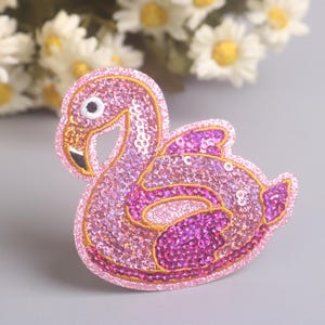 Könnte beinhalten: Ein pinkfarbener Pailletten-Flamingo-Aufnäher mit goldenem Umriss. Der Flamingo hat einen schwarzen Schnabel und ein schwarzes Auge, vor grauem Hintergrund mit weißen Blumen.