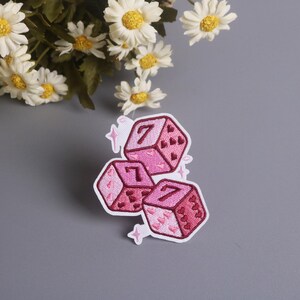 K&ouml;nnte beinhalten: Drei rosa W&uuml;rfelaufn&auml;her mit der Zahl 7, bestickt mit Herzen und wei&szlig;en Konturen. Auf grauem Hintergrund, mit wei&szlig;en G&auml;nsebl&uuml;mchen. Ideal zum Aufn&auml;hen auf Kleidung.