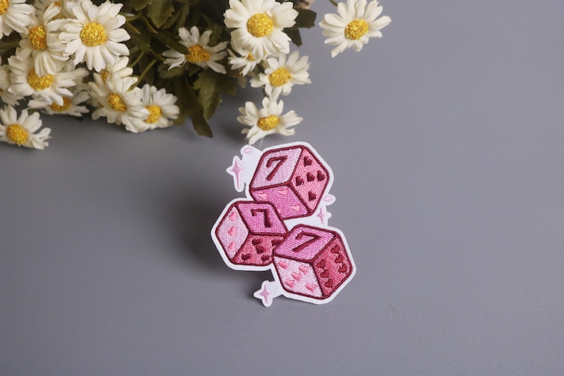 K&ouml;nnte beinhalten: Drei rosa W&uuml;rfelaufn&auml;her mit der Zahl 7, bestickt mit Herzen und wei&szlig;en Konturen. Auf grauem Hintergrund, mit wei&szlig;en G&auml;nsebl&uuml;mchen. Ideal zum Aufn&auml;hen auf Kleidung.