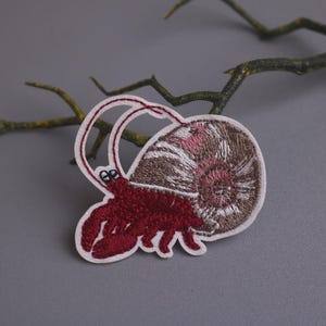 May include: Embroidered patch of a red hermit crab carrying a brown and white seashell on its back.