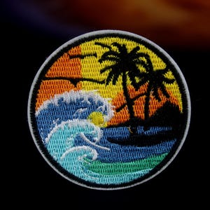 Pode incluir: Um remendo redondo bordado com uma cena de pôr do sol colorida que apresenta duas palmeiras, uma onda e um sol. O remendo é branco com um contorno preto.